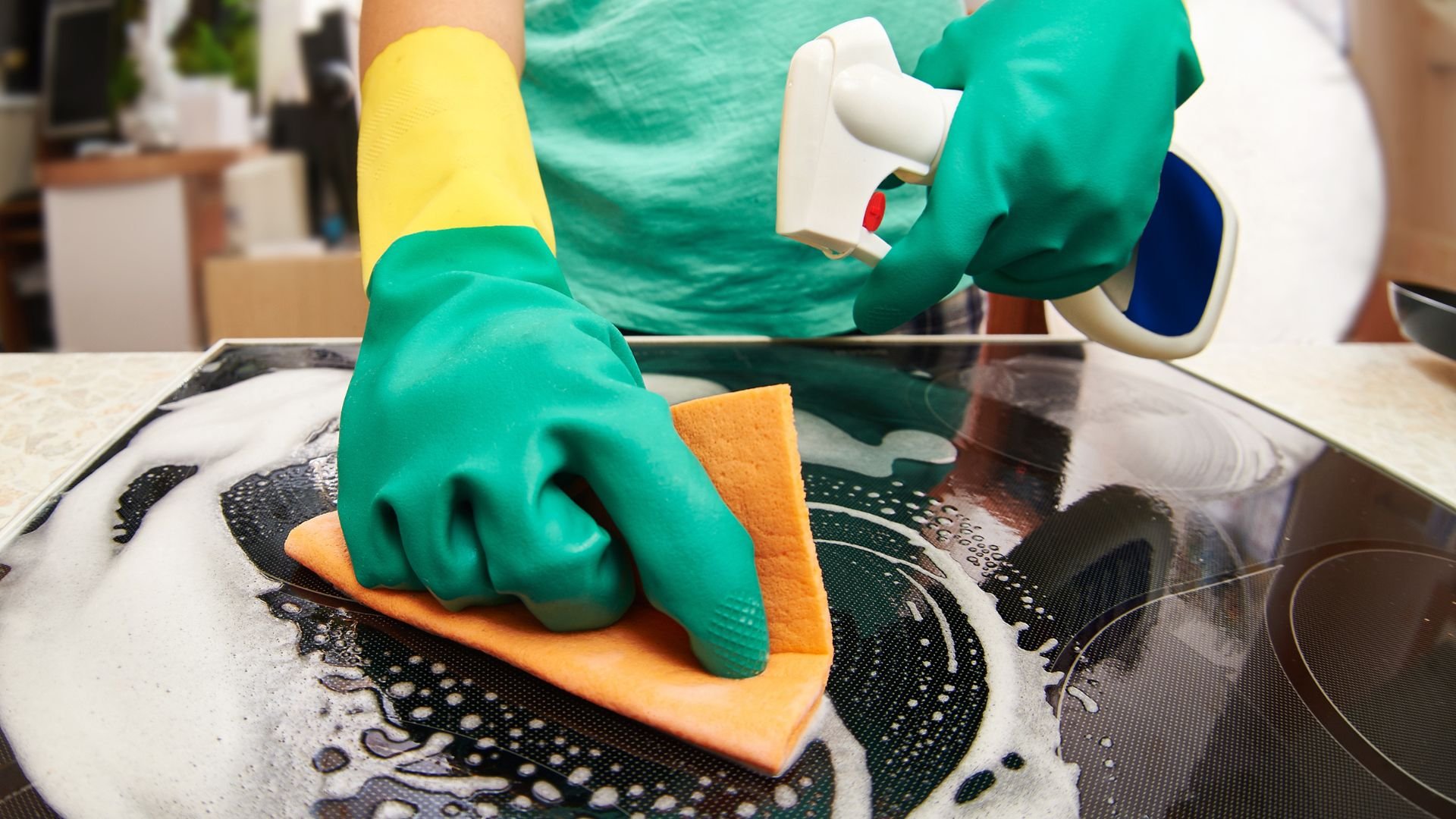 Green gloved hands cleaning stovetop with orange sponge and spray cleaner