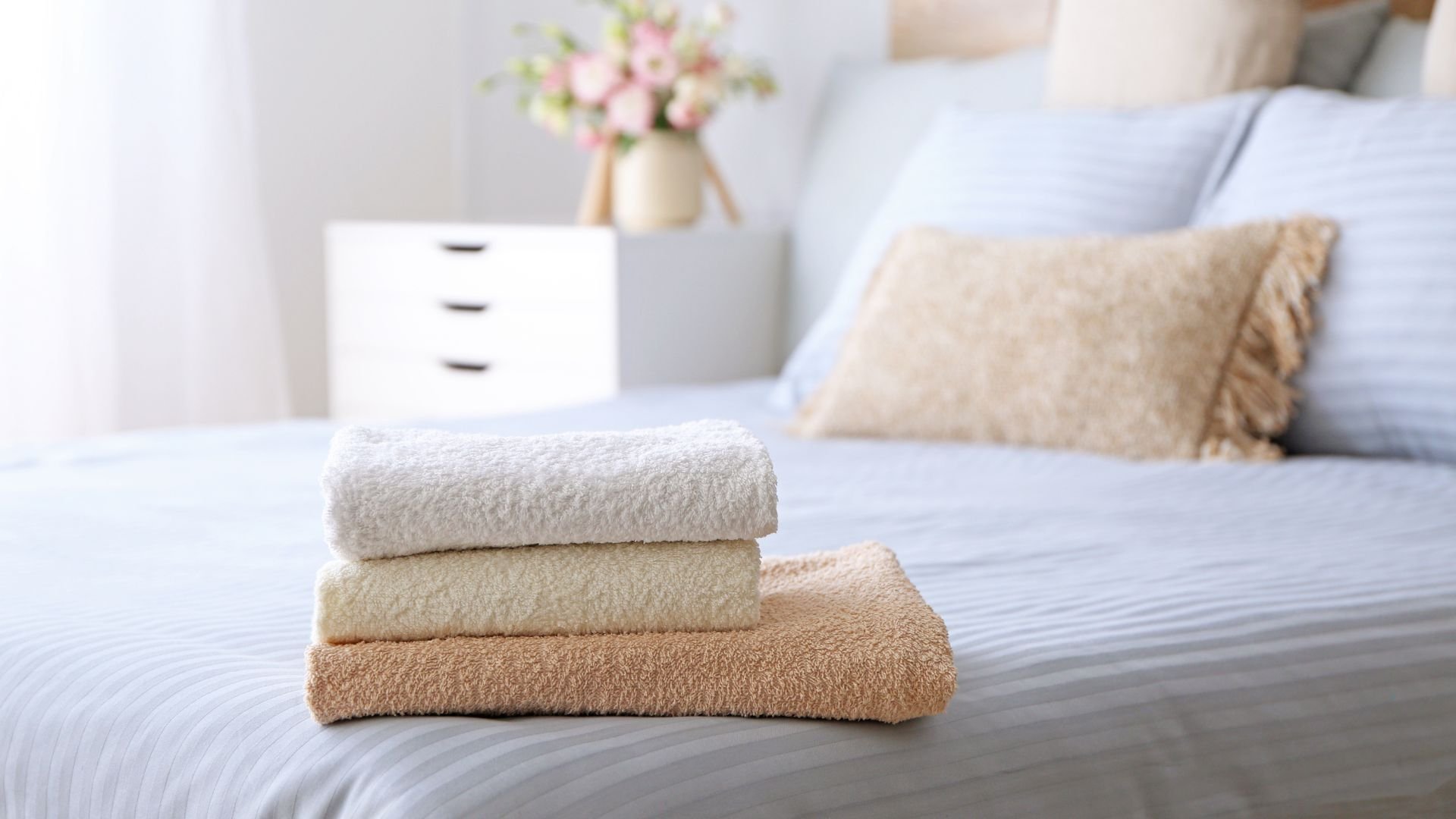 Three soft towels stacked on bed with neutral-colored bedroom background