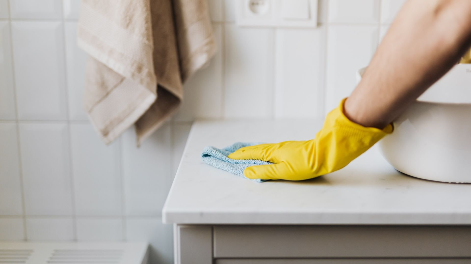 Yellow gloved hand cleaning white surface with blue microfiber cloth